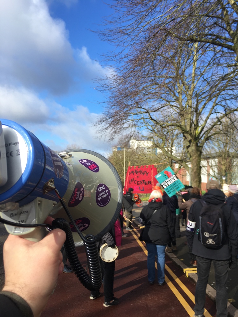 Point of view of a person carrying a megaphone covered in UCU stickers, following a group of striking colleagues on a march who are carrying banners and UCU signs.
