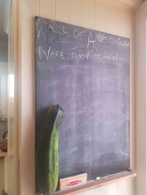 Black blackboard, located in a kitchen of a home-working staff member, reading "Wall of Appreciation Near the Kitchen" written in chalk. Accompanied by an enormous home-grown courgette. 